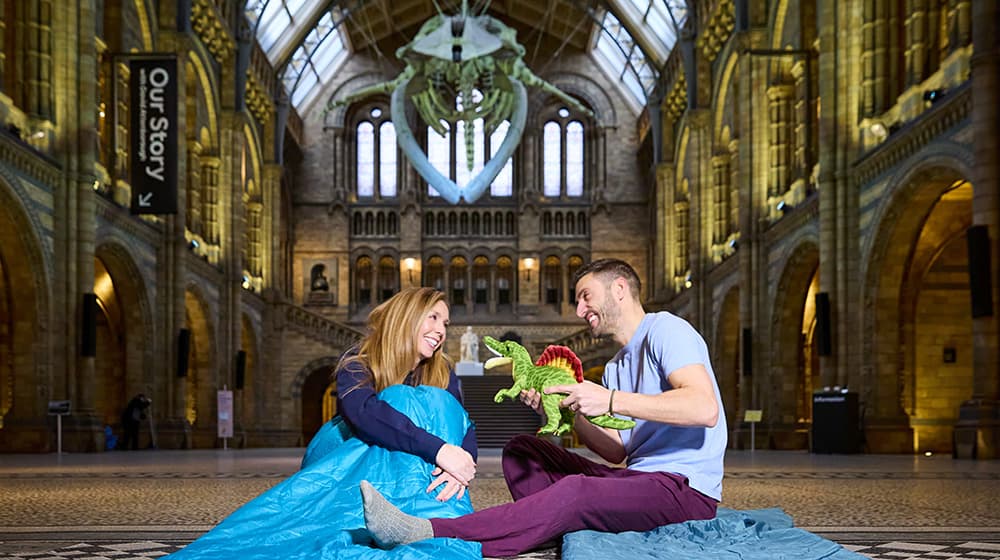 A man holds a dinosaur stuffed toy, sitting next to a woman on sleeping bags in Hintze Hall under Hope the blue whale.