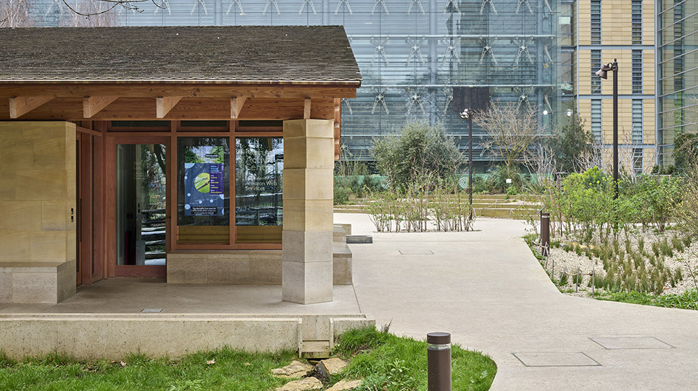 Side on view of the Nature Activity Centre. A beige building with a wooden panelled roof. The Building is surrounded by grassy areas, trees, a pavement area and the glass exterior of the Museum can be seen in the background.