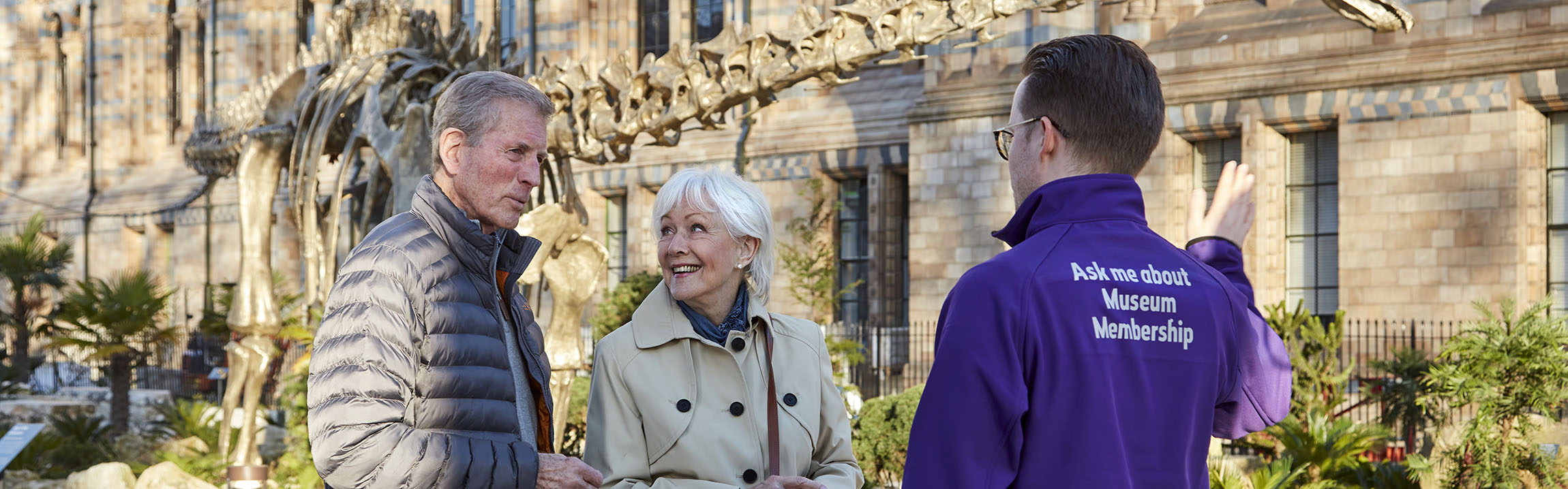 A guide talks to two visitors at the Museum gardens with Fern the Diplodocus in the background.