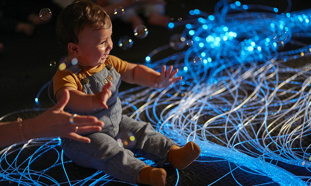 A baby sits sits in front of blue lighting as an extended hand helps him