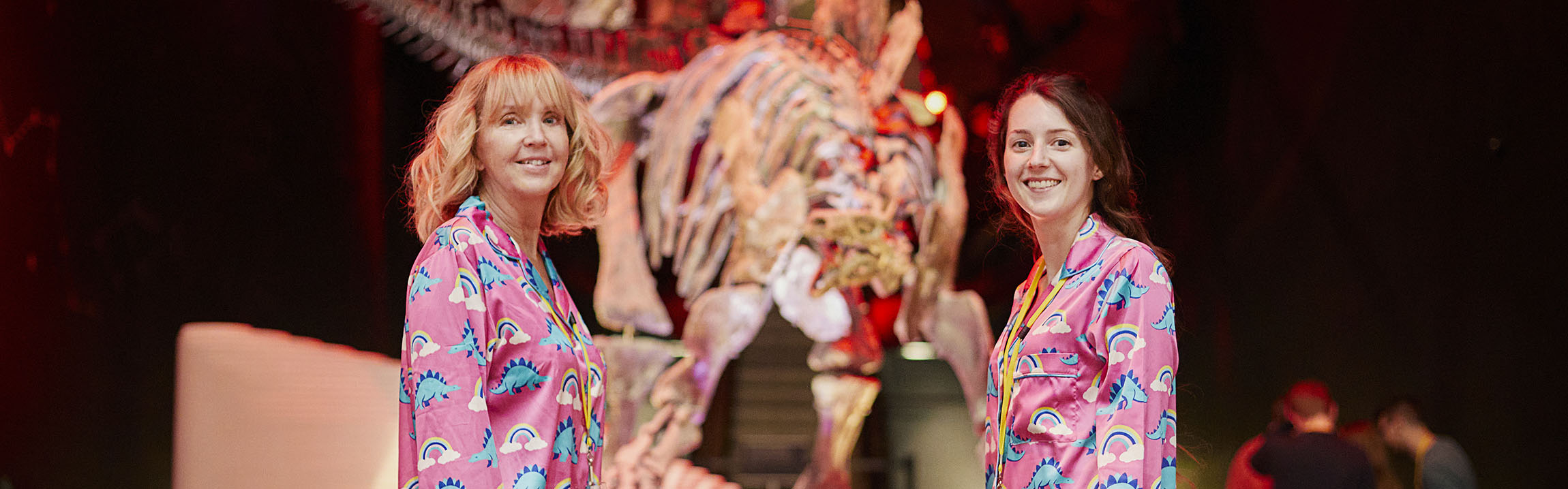 Two women wearing pink pijamas with dinosaurs on them pose in front of Sophie the Stegosaurus