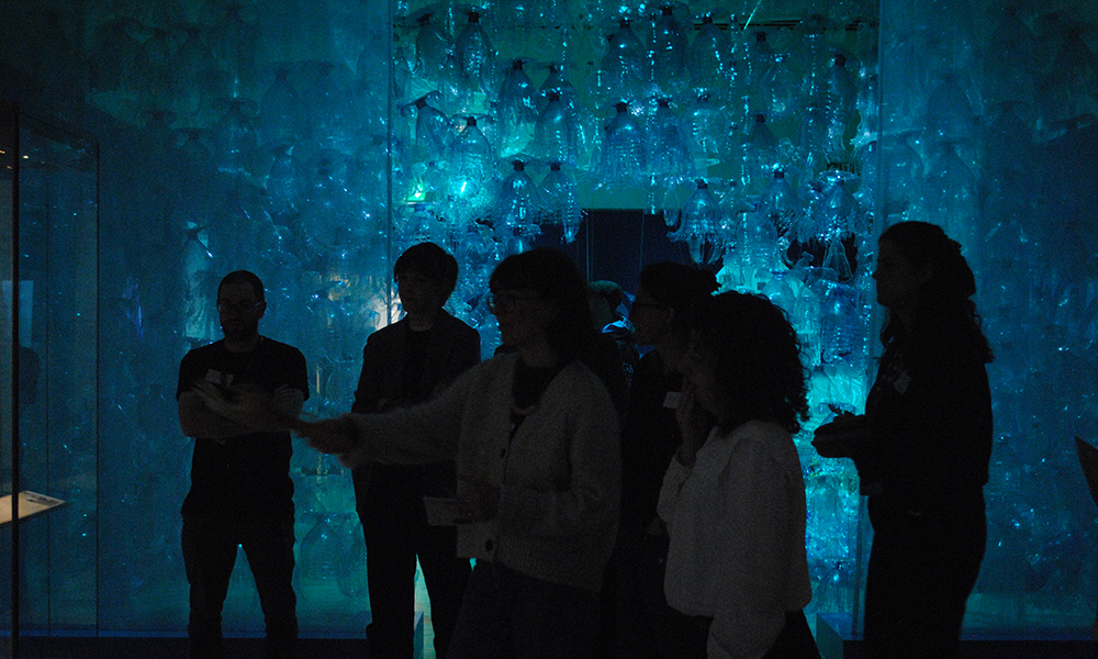 A photograph of a group of people standing in a dark room. Their silhouettes are lit by a wall of blue plastic bottles lit in blue light.