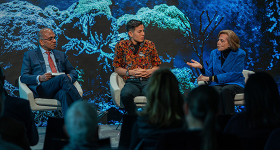 Three speakers are seated on a stage in front of a projected image of a coral reef