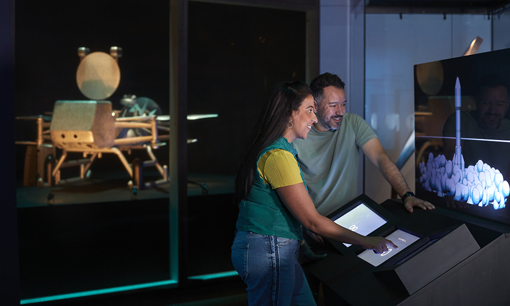 A man and a woman touch a screen as another screen shows them a white rocket launching