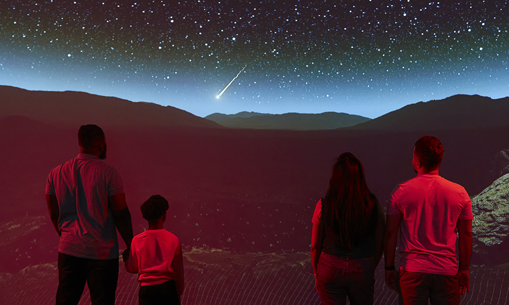 Four people look at a projection of Mars on a big screen, with with red soil and the night sky full of stars 