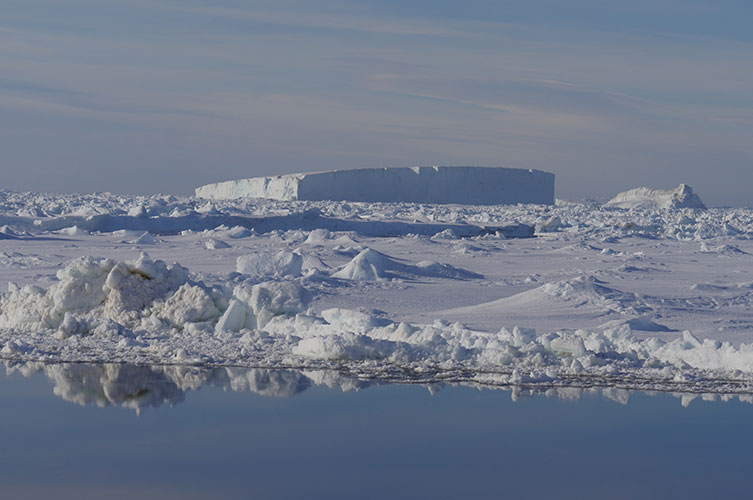 An expanse of ice and snow beside water. A tall, cubic iceberg is visible in the distance. 