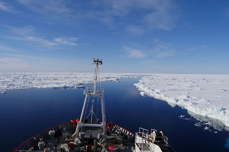A large boat travels through a calm channel of water between large, flat patches of sea ice. 