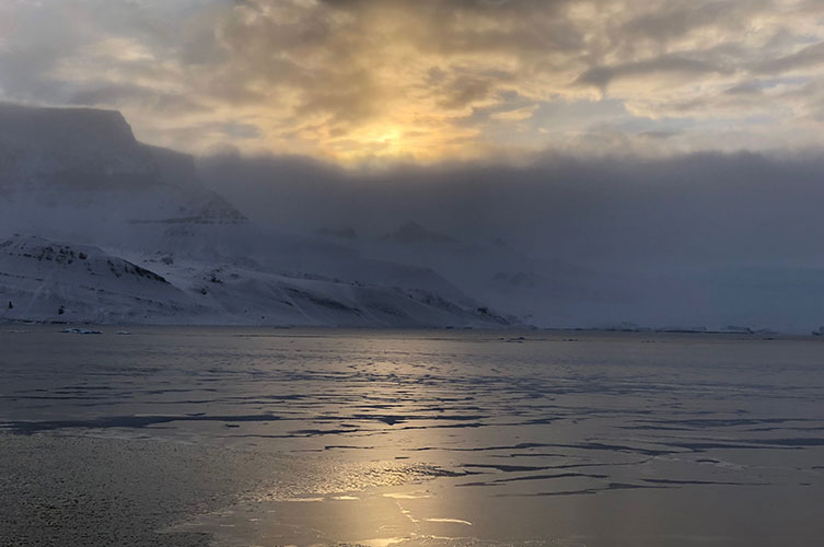 Sunlight reflects off ice on the ocean's surface next to tall, snow-covered mountains on Antarctica's coast. 