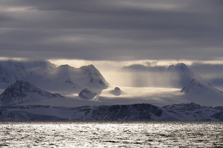 Sunbeams fall on tall, snow-covered Antarctic mountains, close to the coast. 