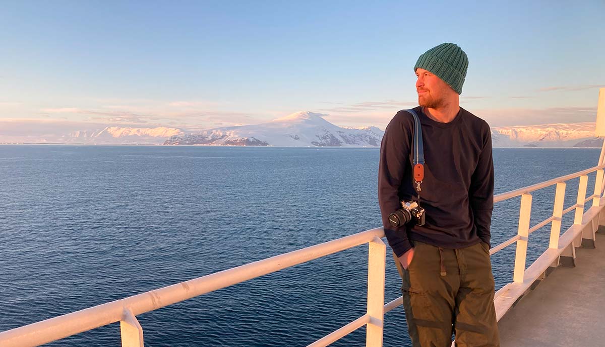 Dr Hugh Carter wearing a woolly hat and jumper leans against the railing of a ship with a snow-capped mountain in the distance.