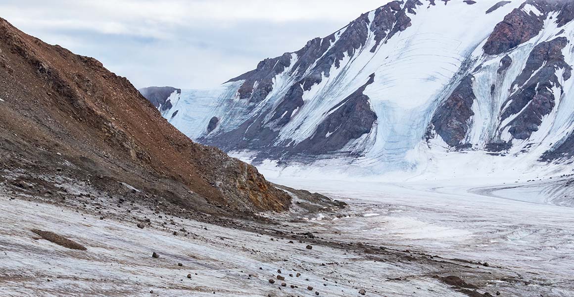Bare rocky slopes rise from a glacier in the foreground, with snowy mountains rising in the background. 