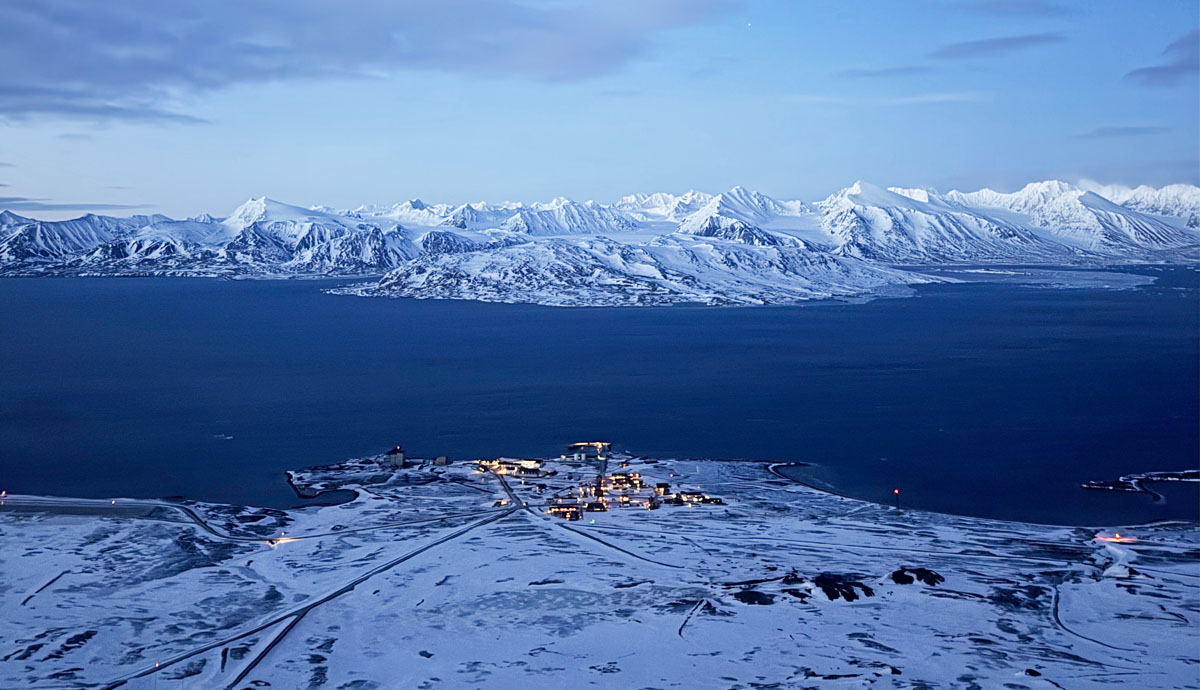 A wide view of the town of Ny-Ålesund on land next to water. In the distance are snow covered mountains.