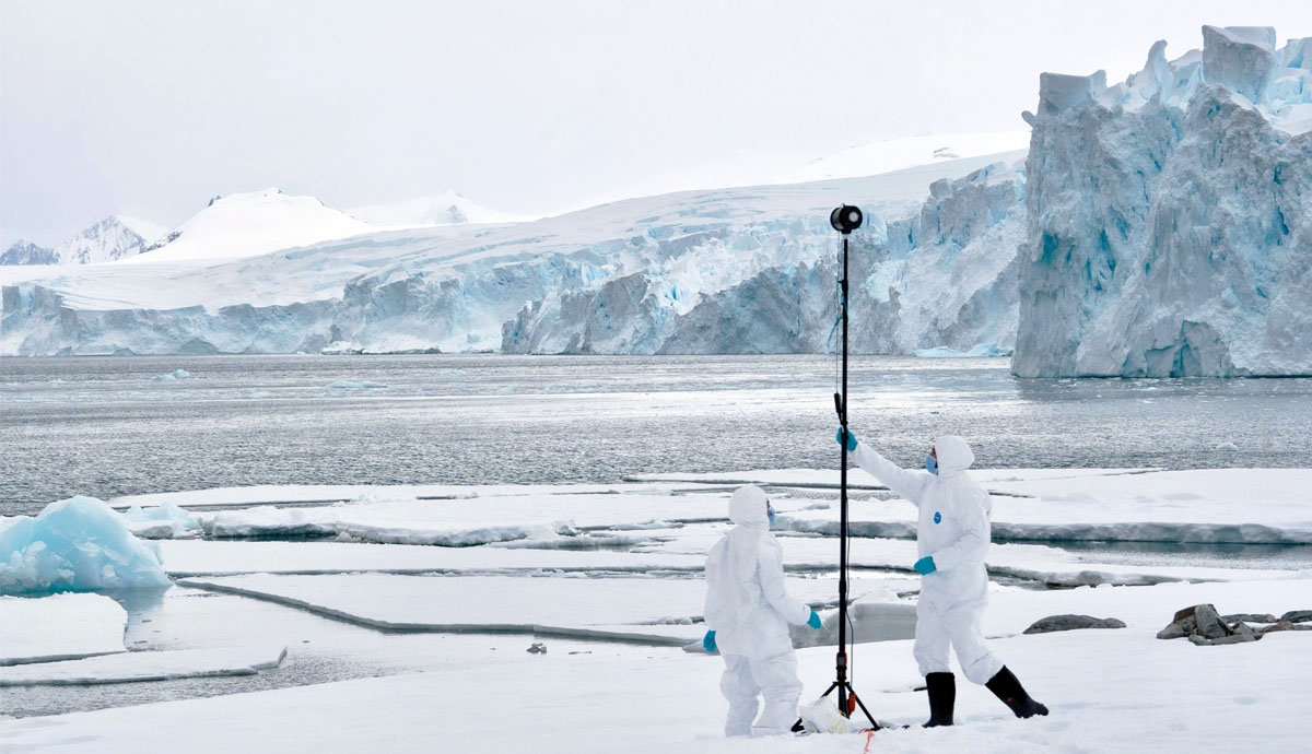 Two scientists in white overalls and a facemask surounded by icy cliffs. They are operating a tall instrument used for air sampling.