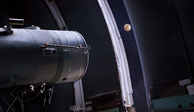 A large telescope looks at the Moon through a gap in an observatory roof.