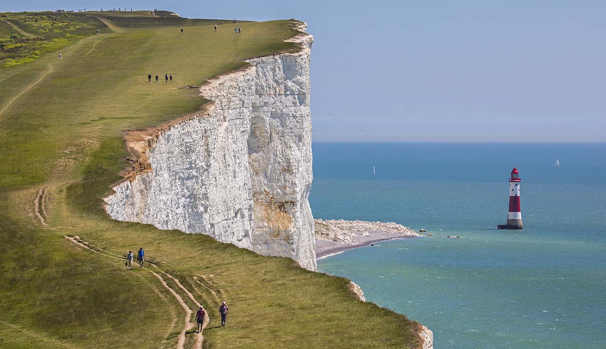 A photo of walkers taking a path along the cliffs at Beachy Head, with the red and white striped lighthouse seen just beyond the shore.