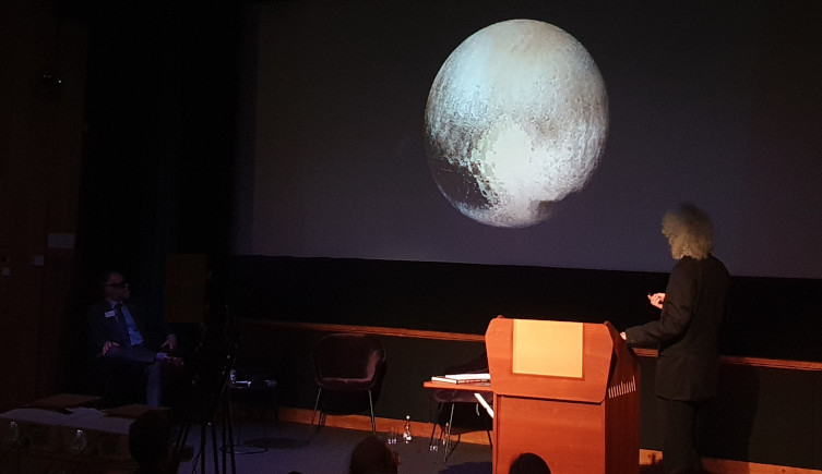 Dr Brian May looks towards a stereoscopic image of Pluto being projected on a screen during a presentation.