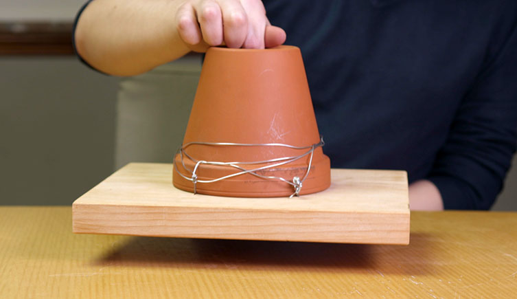 A person lifts the pot, showing that it is securely attached to the piece of wood