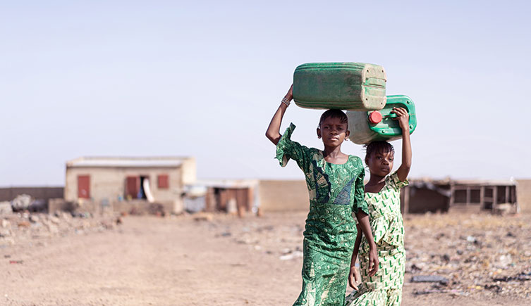 Two girls carry water containers on their heads as they walk down a dusty path