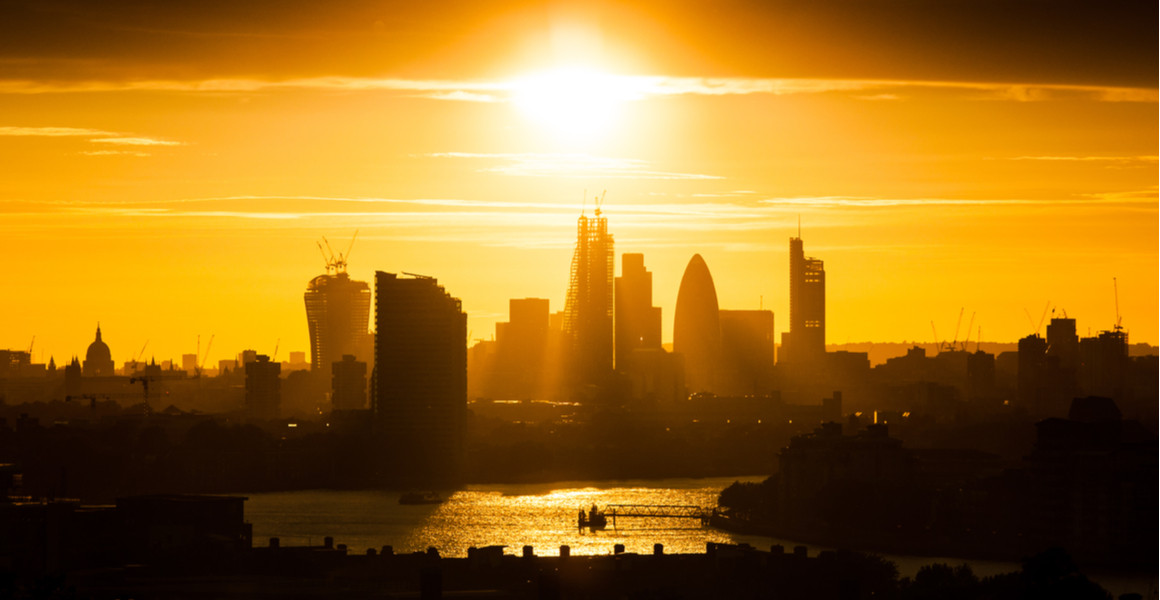 The sun hangs over London, silhouetting tall buildings such as the Gherkin