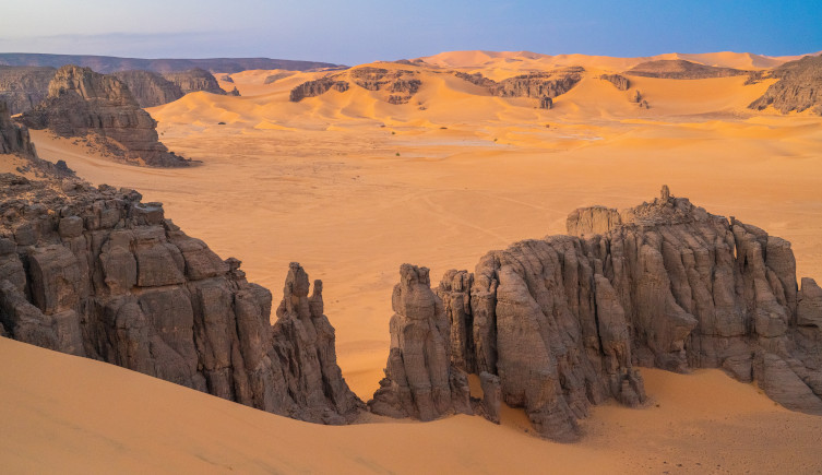 Rocks rise out of the sand in the Sahara Desert, with mountains in the distance.