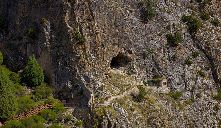 A photograph of a cave opening in the side of a sheer mountain, with a building nearby.