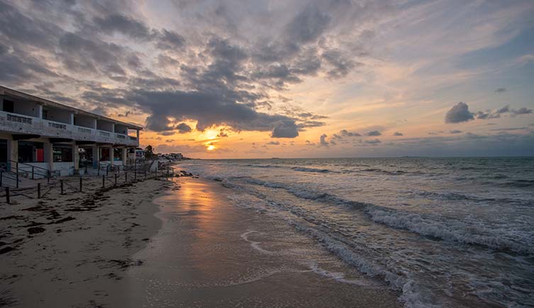The sun sets over a beach, with a blocky building on the coast.