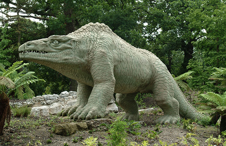 A green, four-legged sculpture of a large lizard with sharp teeth surrounded by plants.
