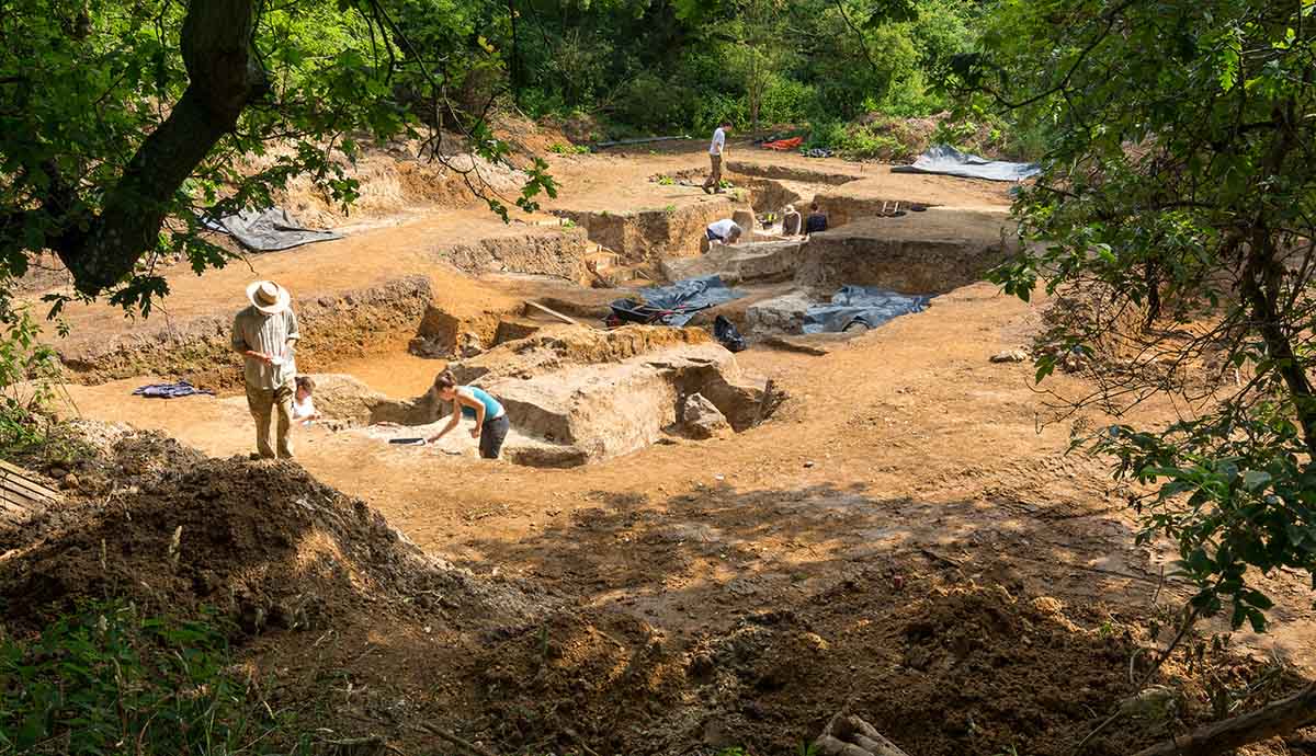 A view looking down into the Barnham site, showing a large bare patch of ground pockmarked with holes surrounded by trees and greenery. There are a few people stood around the site.  
