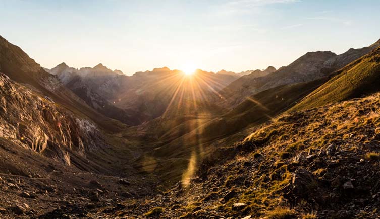 The sun shines through a valley as it peeks out over a mountain range.