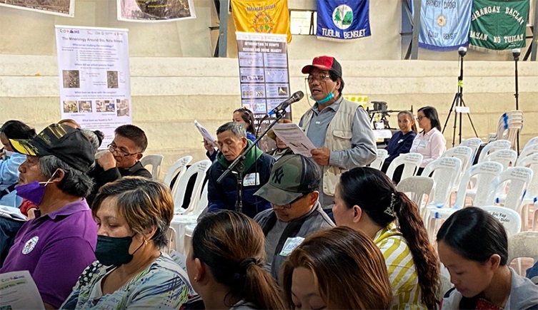 A community meeting in which about 10 people of varying ages are sat in rows on plastic chairs. One person wearing a cap and glasses is stood up in front of a microphone and looks like they are reading from a piece of paper they are holding in their hands. 