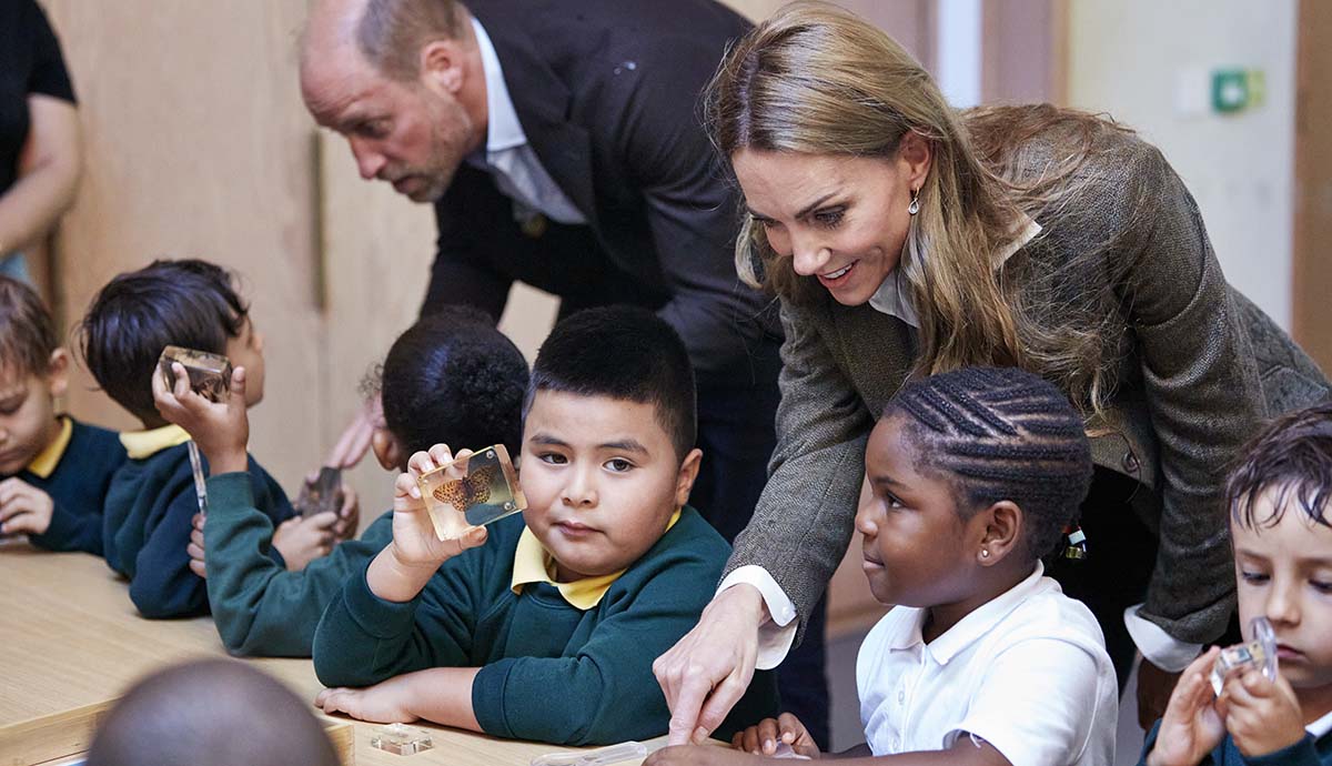The Prince and Princess of Wales leaning over school children helping them to do an activity. 