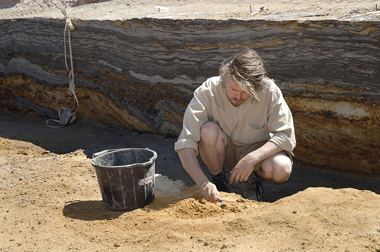 Person crouched down in an excavation pit, using a trowel to sift through sediment.