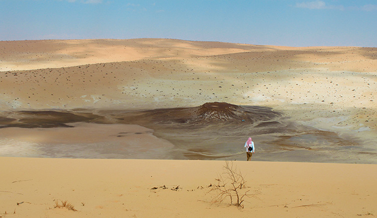 A figure stands on the crest of an orange sanddune, looking down into the space created between two dunes. 