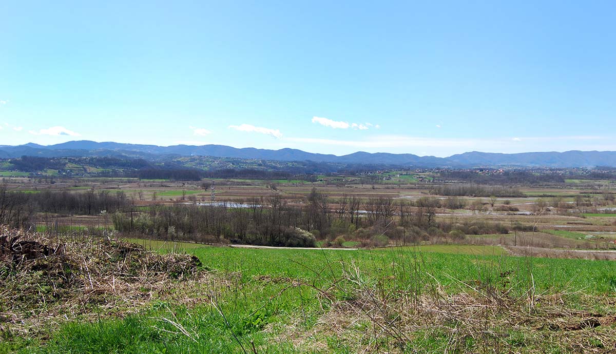 A view across a river running through farmland, with mountains rising in the background.