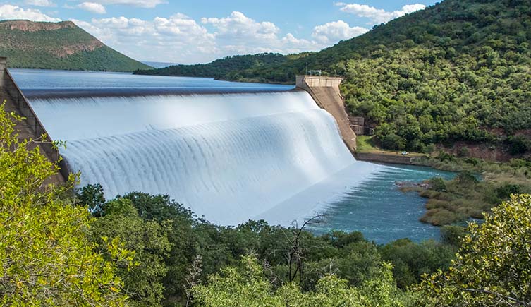 A massive weir with water flowing over it, set amongst a forested landscape. 