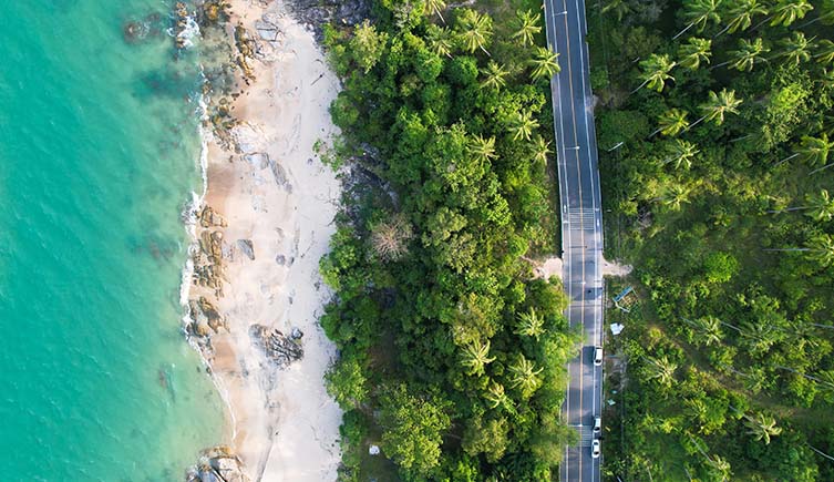 An aerial view of a beach next to a tropical forest with a cutting through it. 