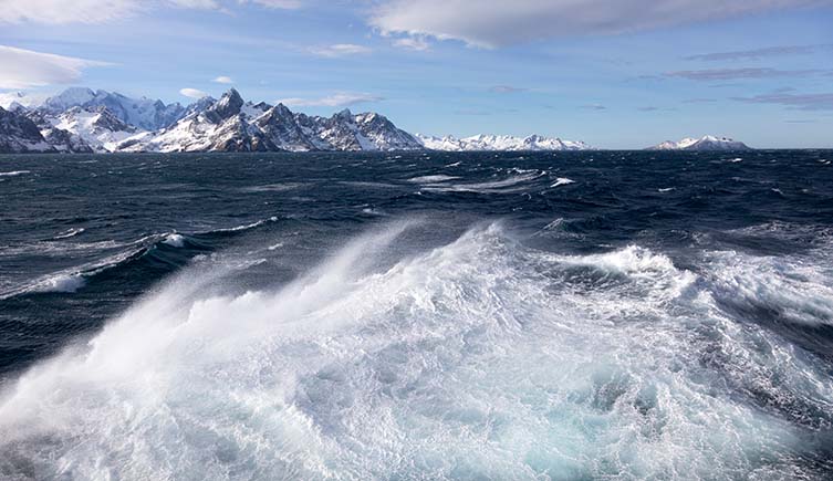 Waves on the surface of the sea with snowy mountains in the distance.