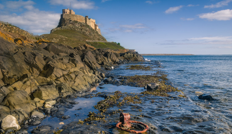 Lindisfarne castle sits on a hill overlooking a rocky coastline, with a rusty iron ring in the sea