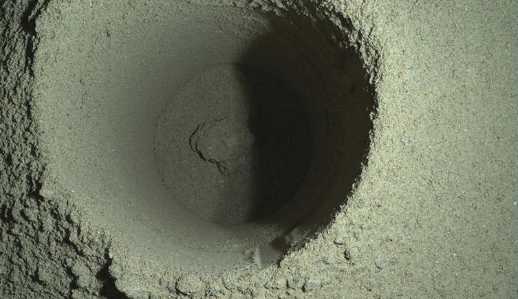 A close-up photograph looking straight-down of a circular hole drilled into the surface of Mars. The soil looks sandy.  