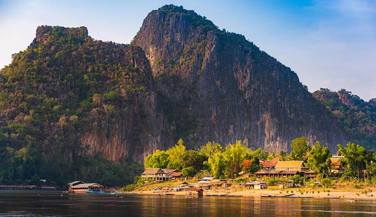 Sheer rocky mountains rise up behind a village on the banks of the Mekong River.