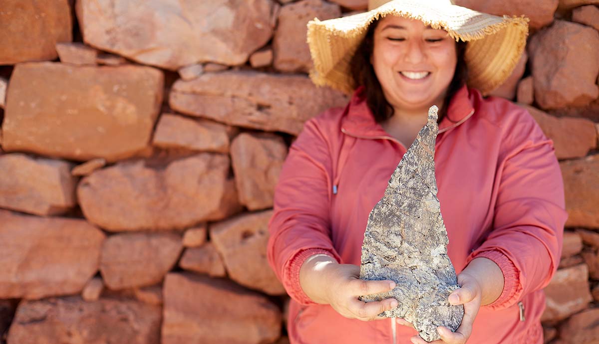 A woman in a floppy straw hat standing in front of a stone wall holds a large fossil spike.