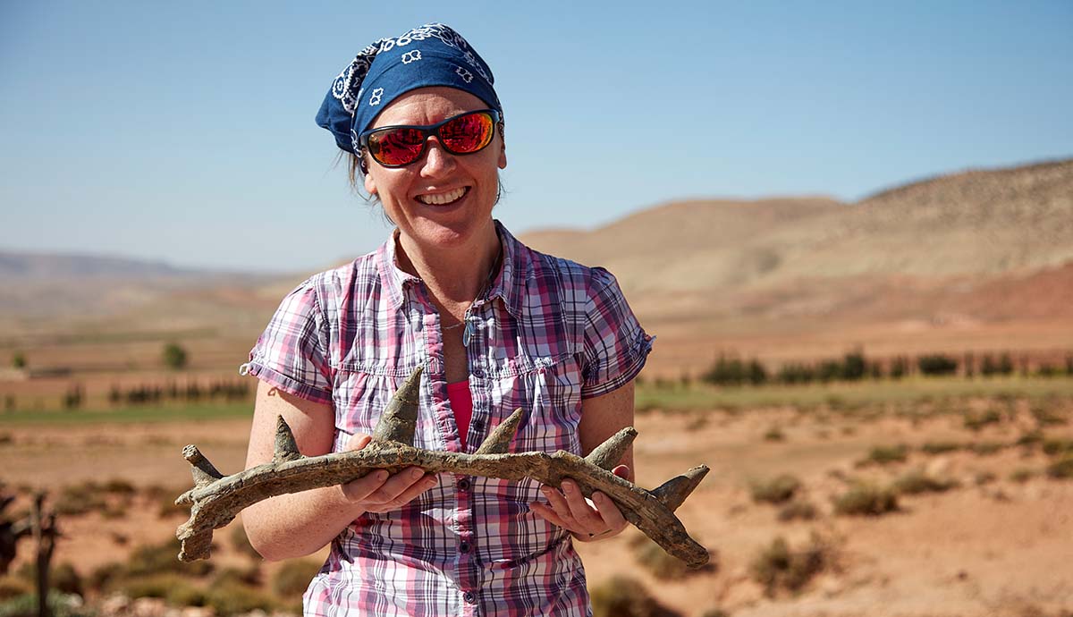 Professor Susannah Maidment, wearing sunglasses and a bandana, holds a bone with several spikes emerging from it.