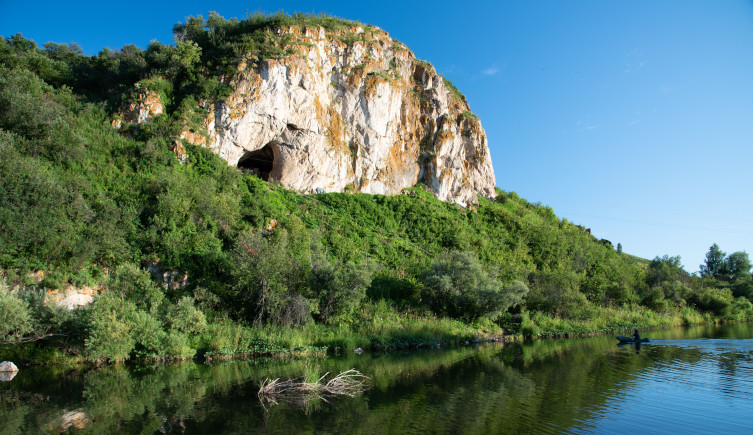 River passes by a rocky outcrop in woodland, which contains Chagyrskaya cave