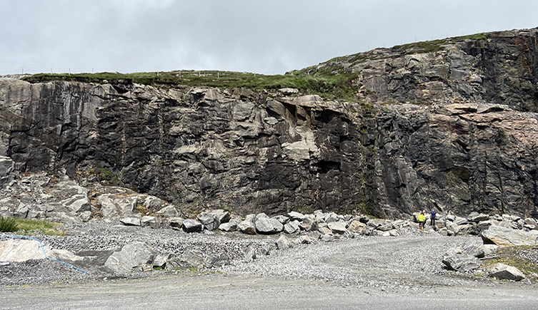 A rock face that was once a quarry with boulders sitting at its base - the boulders are about the same size as two people standing at the quarry base 