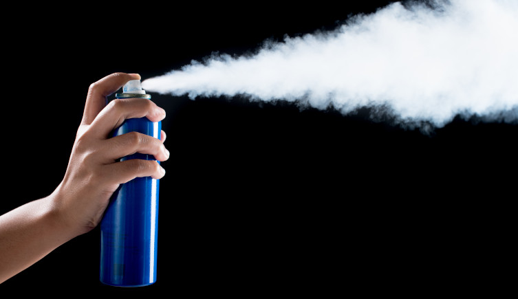 An aerosol can held in a hand sprays white gas against a black background.
