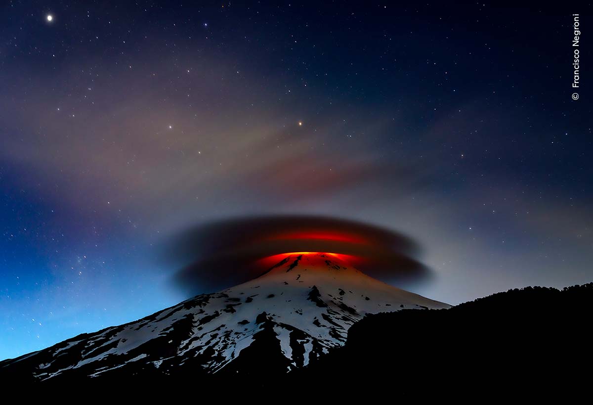 A snowcapped volcano with two glowing lenticular clouds hanging above it beneath a starry sky.