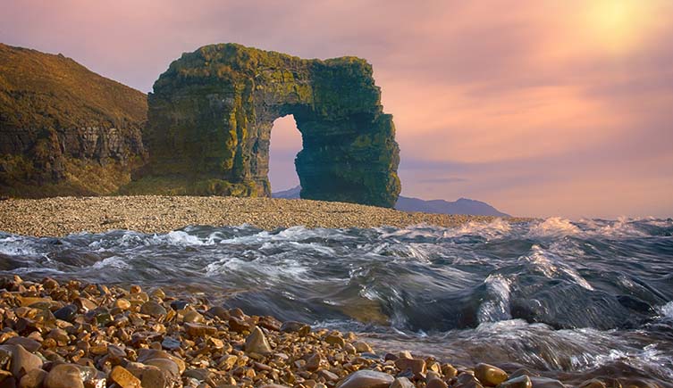 Steller's Arch, a natural rock formation on Bering island, stands on a beach behind waves in the foreground. 