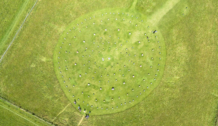 Looking directly down on woodhenge, revealing a circular pattern of grey stones set against the green grass. 