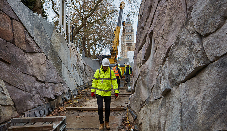 A person in working gear and a hard hat walks through a passage with rock walls - the walls and floor are under construction. Trees and the Natural History Museum building are visible at the end of the passage.