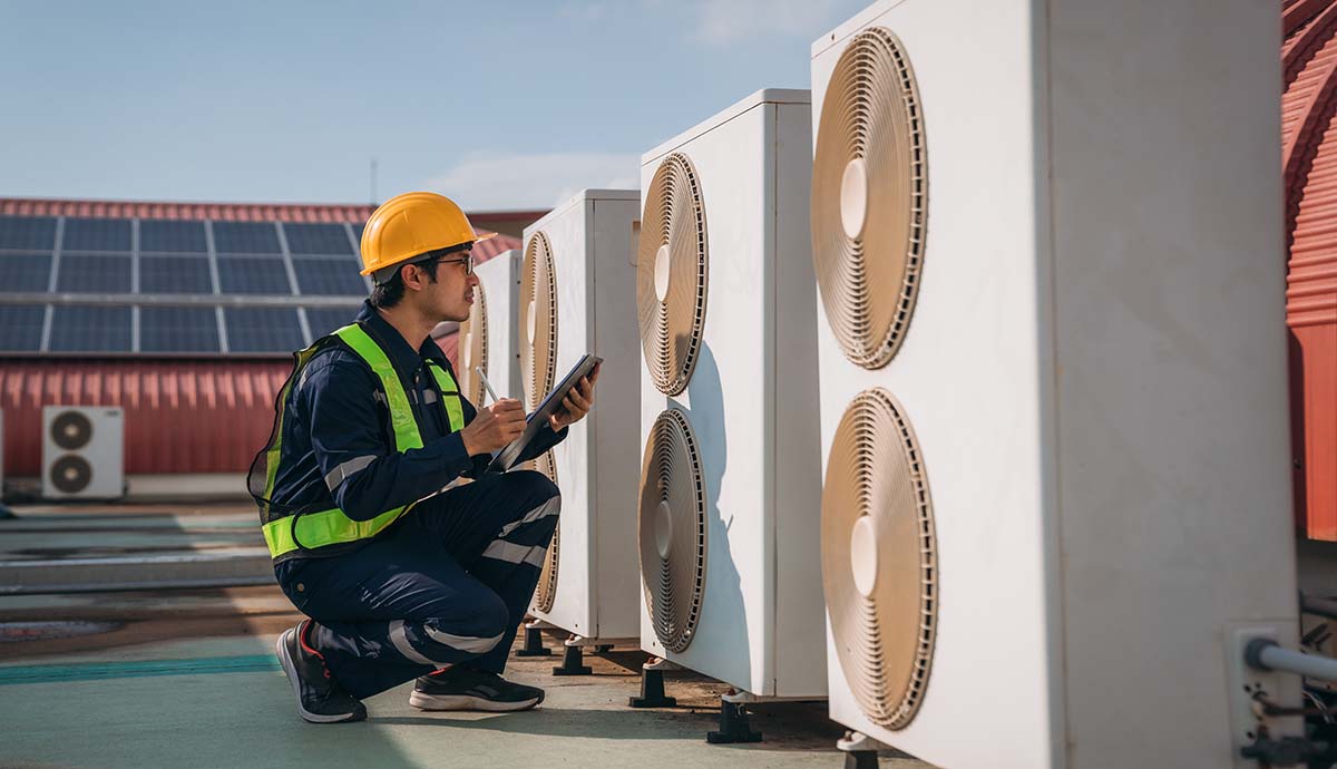 A worker inspects a row of heat pumps on the roof of a building.